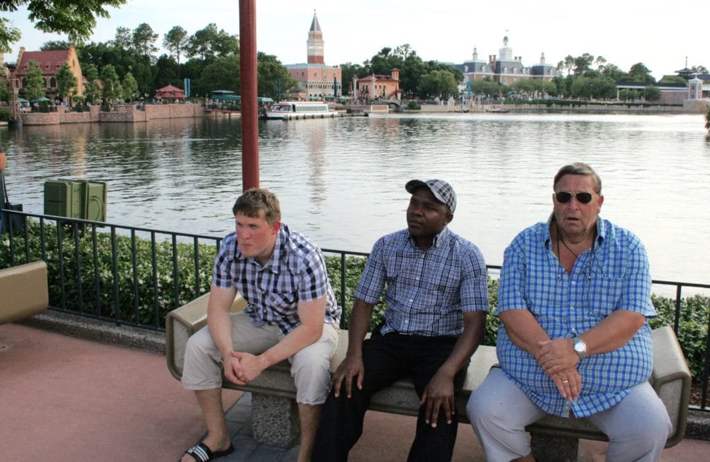 Tre menn sitter på en benk ved vannkanten med en vakker bylandskap i bakgrunnen.