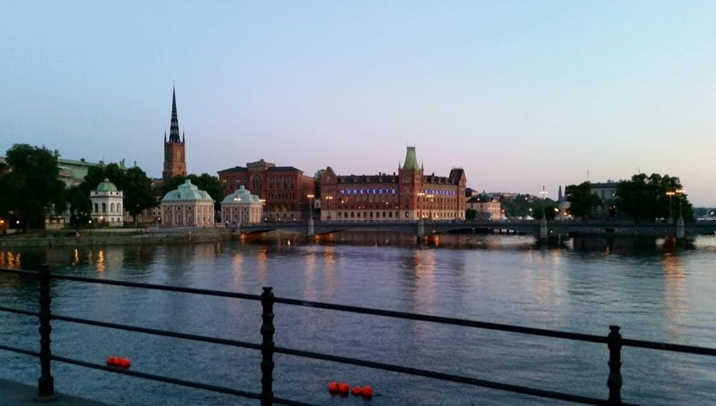 Solnedgang over Stockholms skjærgård med historiske bygninger og en kirkespir ved vannkanten.