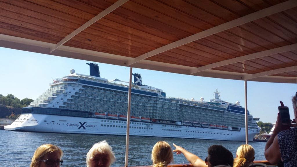 Et stort passasjerskip seiler langs kysten i Stockholms skjærgård, med turister som ser på fra en mindre båt i forgrunnen.