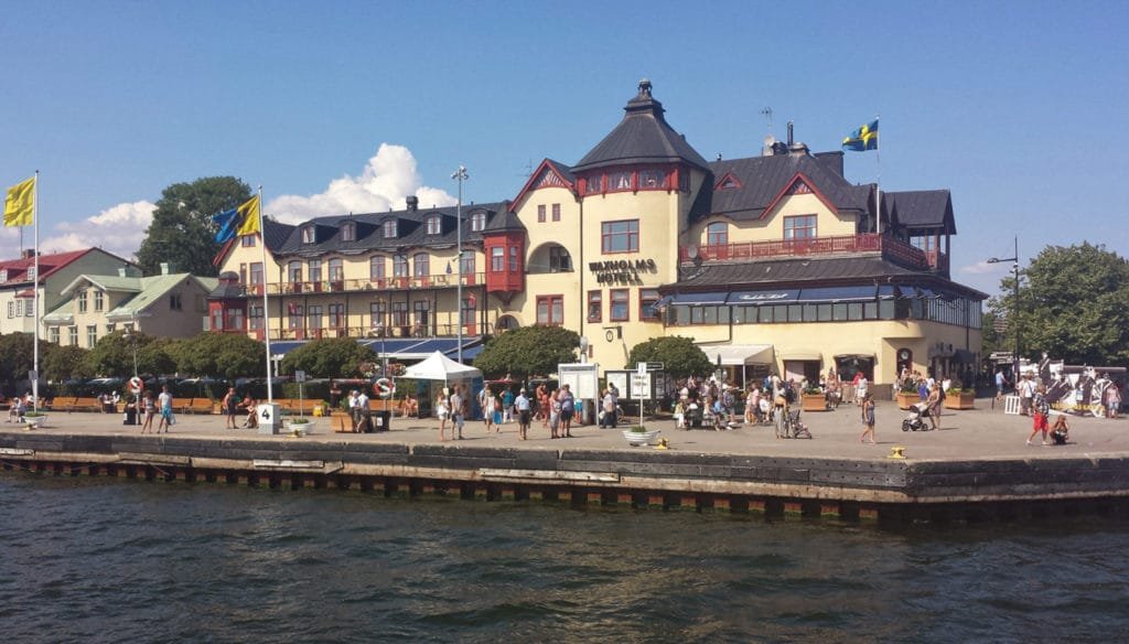 Utsikt over Stockholms skjærgård med et historisk bygg ved vannkanten, utendørs sitteplasser og folk som nyter en solrik dag.