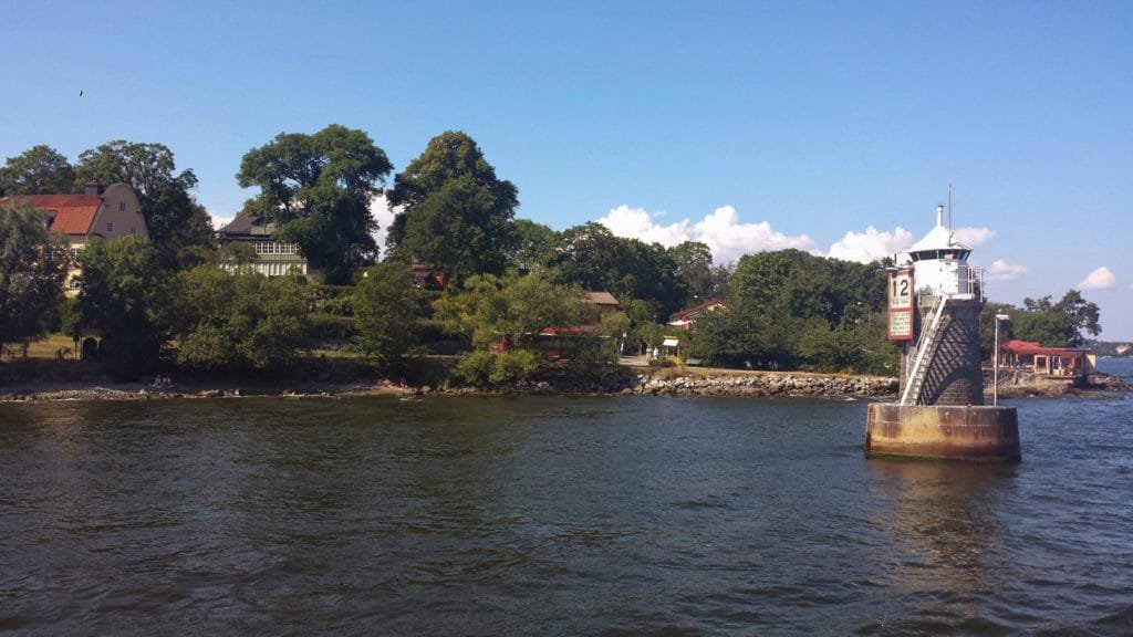 Liten hvit fyr på et grønt skjær med trær og noen hus i Stockholms skjærgård under en klar blå himmel.