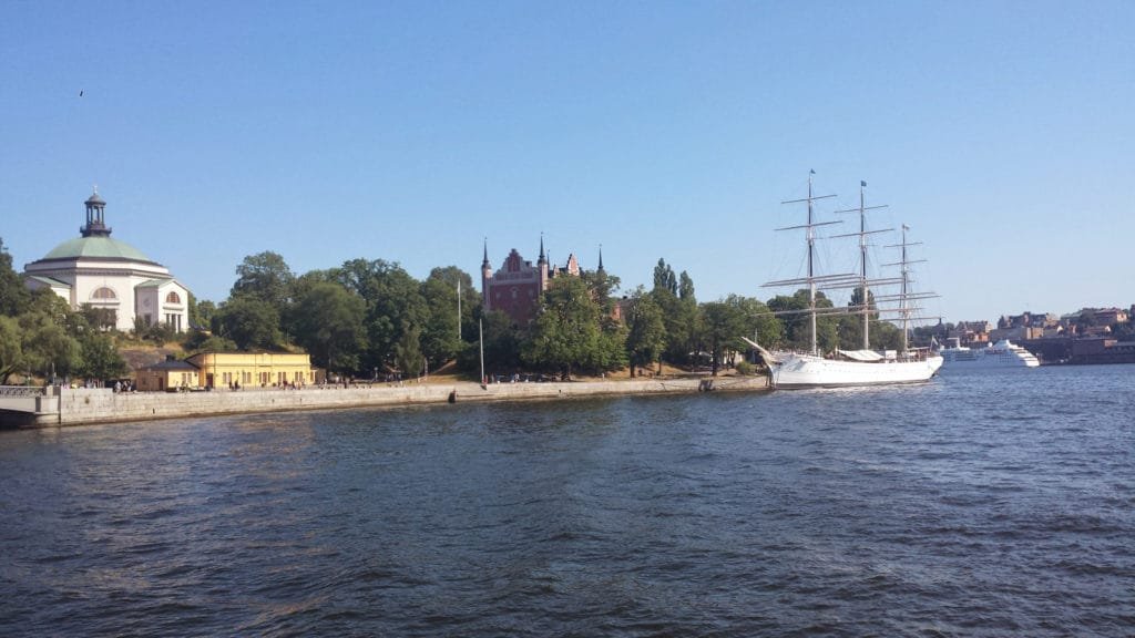 Historisk sjøfront med høyt seilskip og gamle bygninger i Stockholms skjærgård under klar blå himmel.