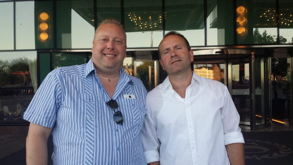 To menn står og smiler utenfor inngangen til en moderne bygning med store glassflater på en solrik sommerdag i Stockholm.