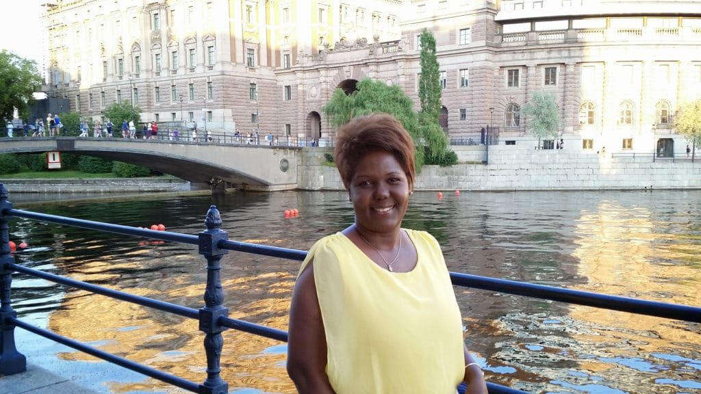 Smilende kvinne i gul ermeløs topp ved elvebredden med rekkverk, med historiske bygninger og bro i bakgrunnen under en solrik sommerdag i Stockholms skjærgård.