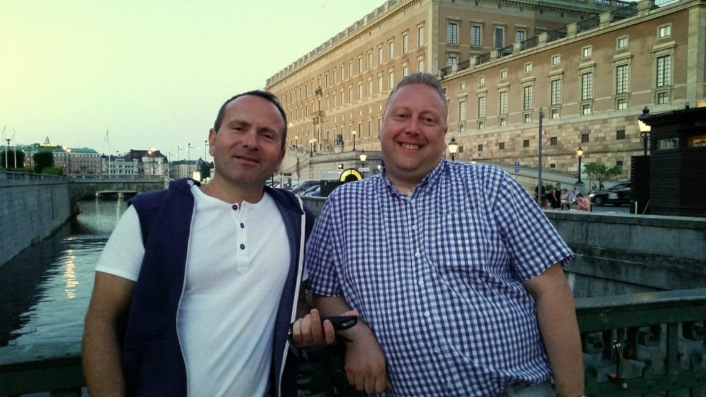 To menn smiler og poserer ved en kanal med historiske bygninger i bakgrunnen.