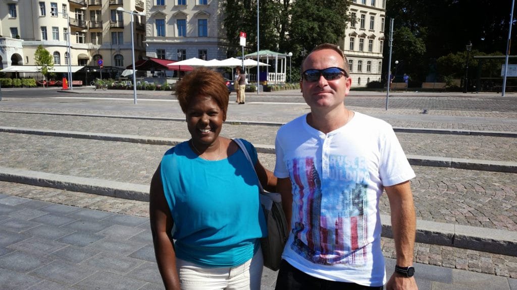 To personer som smiler utendørs i solfylt bymiljø med historiske bygninger i bakgrunnen.