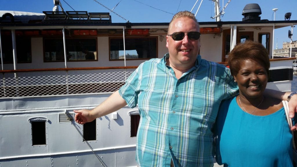 Ett smilende par står foran passasjerbåten S/S Storskär på en solrik sommerdag i Stockholms skjærgård.