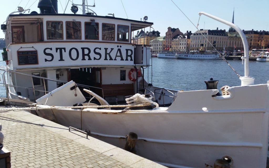 Hvit S/S Storskär-båt liggende til kai i Stockholms skjærgård med bylandskap i bakgrunnen.