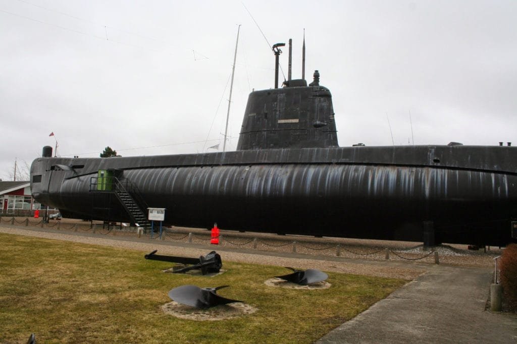 Deaktivert militærubåt utstilt utendørs på et maritimt museum, omgitt av gress og klar himmel.