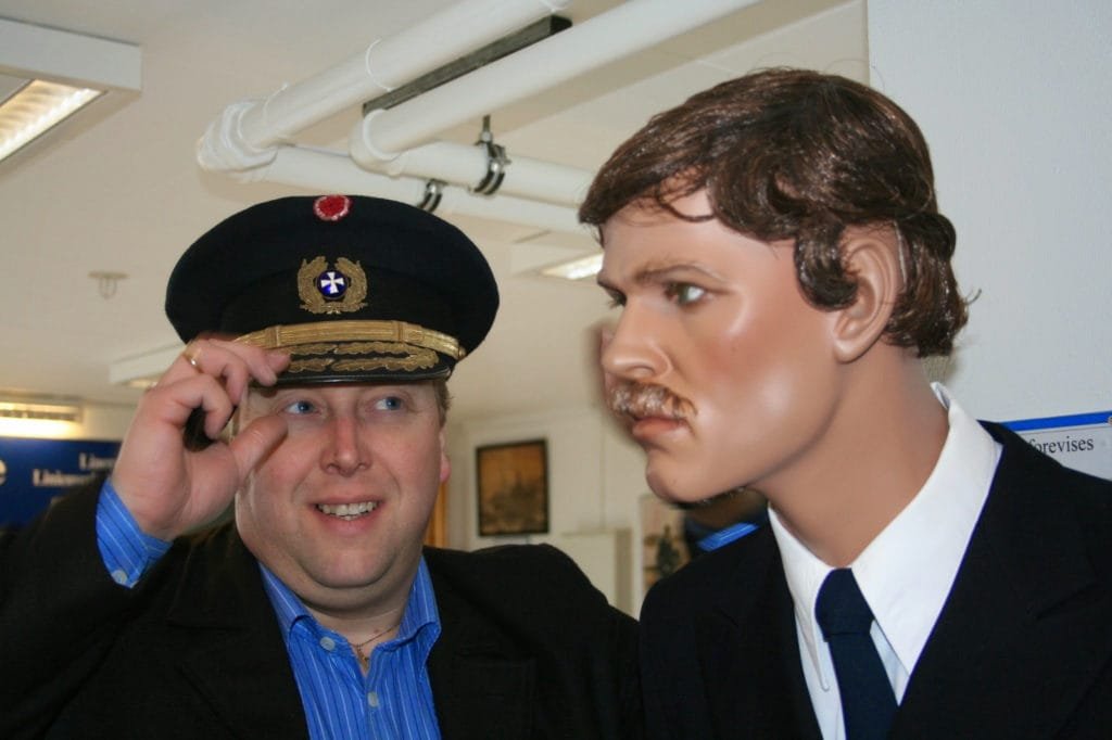 Mann med kapteinlue står ved siden av en livaktig dukke kledd i pilotuniform.