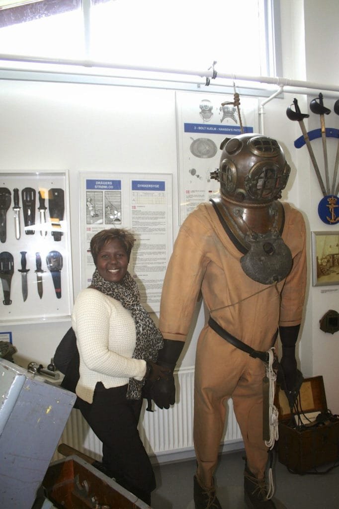 Kvinne poserer ved siden av en vintage dykkerdrakt i et museum.