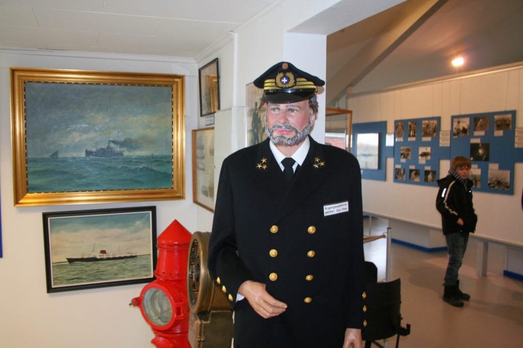 Utstilling på maritimt museum med en ubåtkapteinmankequin i uniform foran historiske malerier av krigsskip.