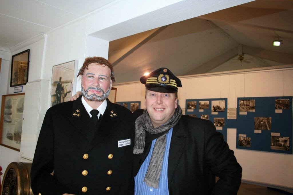 To menn poserer innendørs, hvor den ene er kledd i marineoffisersuniform og den andre har på seg kapteinslue og skjerf.