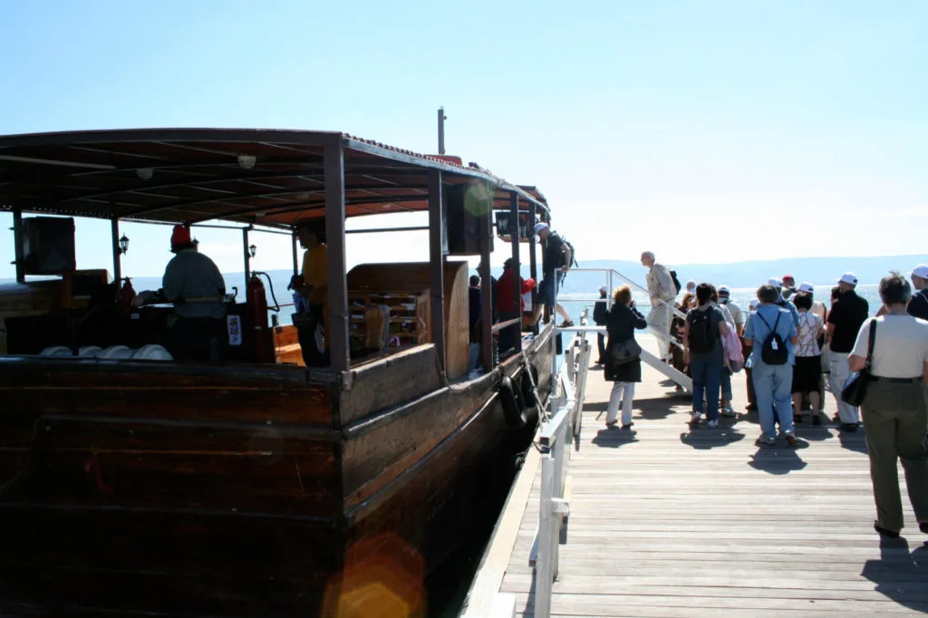 Turister går ombord på en trebåt ved en solfylt brygge ved Genesaretsjøen.