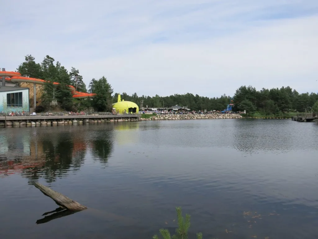 Vakker innsjø med speilblankt vann som reflekterer tettbygde hus og frodige trær langs bredden under en klar himmel.