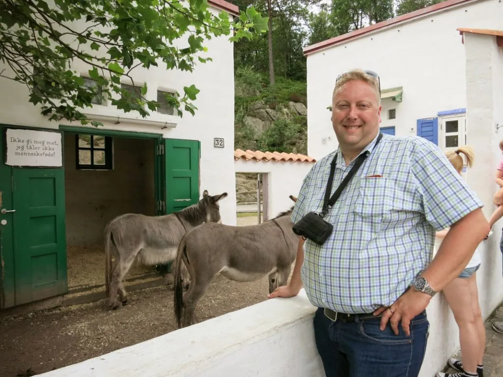 Mann står ved siden av esler foran gårdsbygning med hvite vegger og grønne dører i Kristiansand Dyrepark.