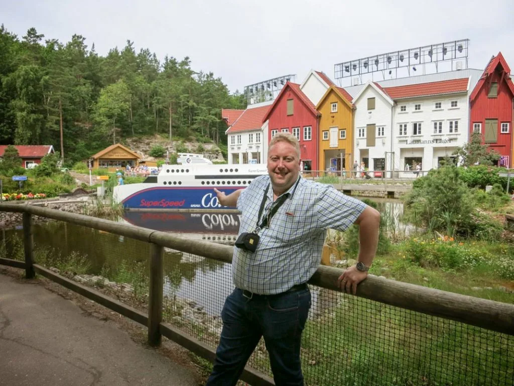 Mann som poserer ved et trestakitt med fargerike bygninger og en båt i bakgrunnen.