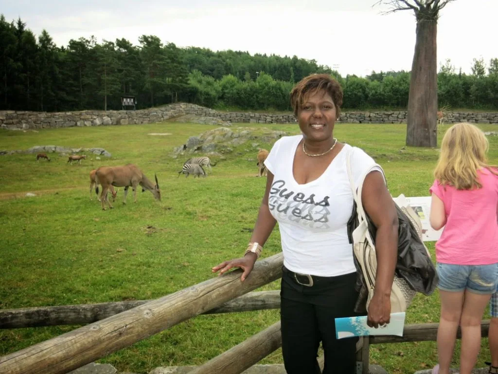 Kvinne som poserer ved et trehekk ved Kristiansand Dyrepark, med dyr som beiter i bakgrunnen.
