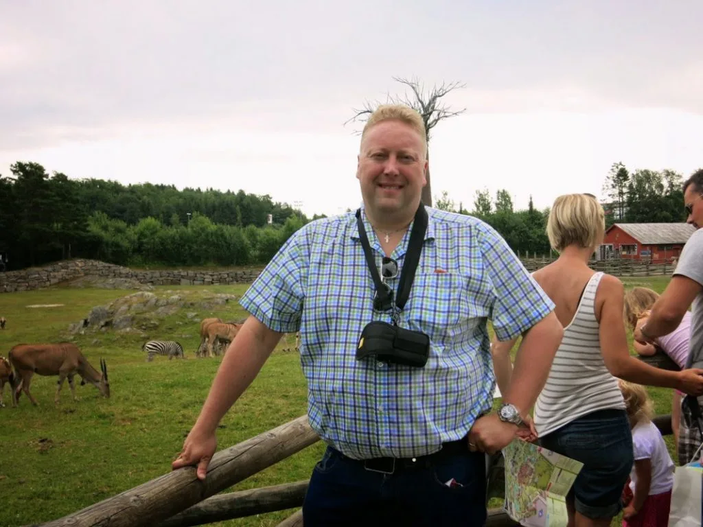 Mann med kamera står ved et gjerde på en bondegård med beitende dyr i bakgrunnen.