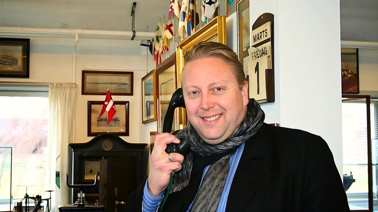 Mann med skjerf som smiler og holder en vintage telefon i et koselig, dekorert rom.